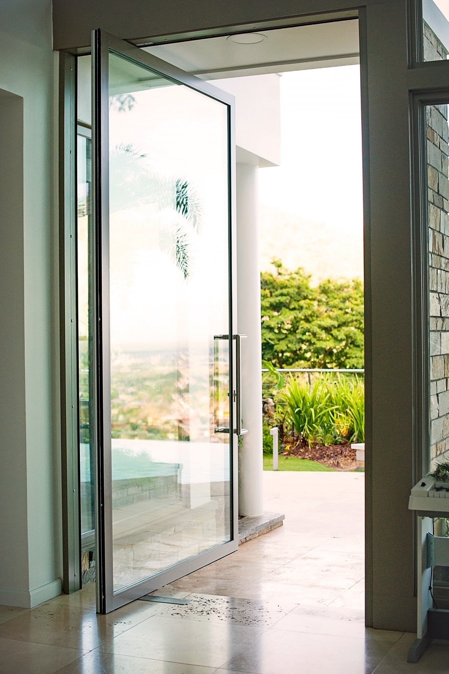 A glass door is open, leading to an outdoor area with lush greenery and partial view of a distant landscape. The interior has light-colored tile flooring.